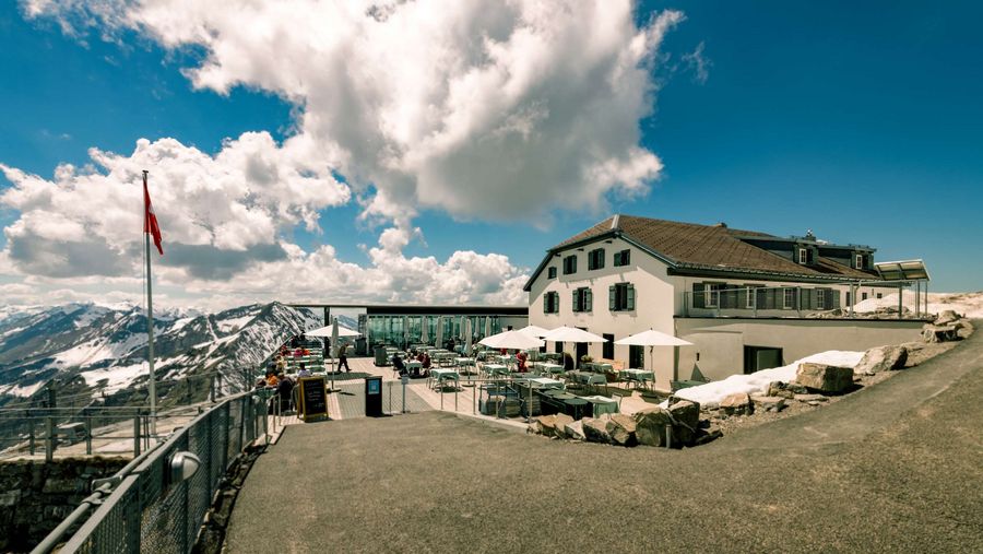 Blick auf das Berghaus Niesen bei sonnigem Wetter, im Hintergrund einige schönwetter Wolken und teilweise verschneite Berggipfel